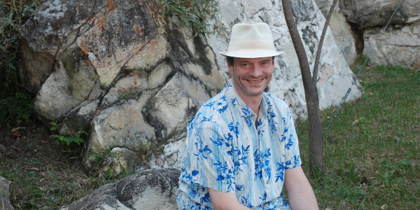 A man wearing a white hat sits on a rock with a rocky nature scene behind him